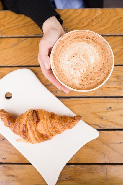 Mano de mujer sosteniendo una taza grande con café capuchino y croissant francés recién hecho en una mesa de madera