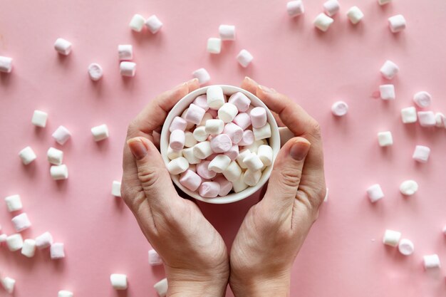 Mano de mujer sosteniendo una taza con mini malvaviscos rosas y blancos sobre fondo rosa