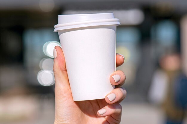 Mano de mujer sosteniendo una taza de papel blanco con café o té con fondo de calle borrosa