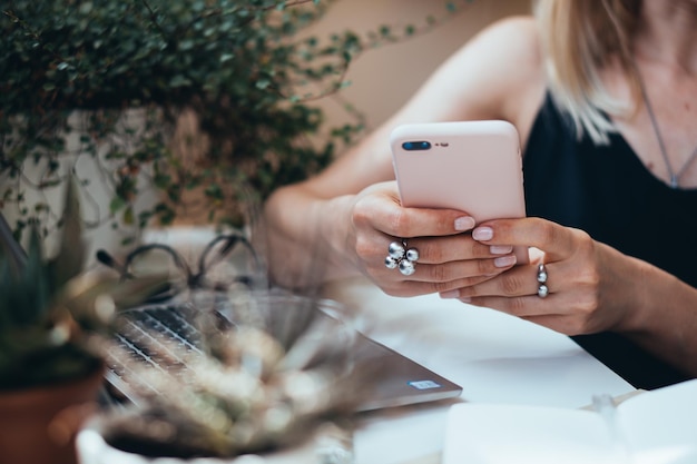 Mano de mujer sosteniendo un teléfono inteligente en una acogedora oficina de verano con computadora portátil y plantas verdes