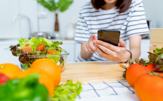 Mano de mujer sosteniendo un teléfono inteligente y una ensaladera con tomate y varias verduras de hoja verde sobre la mesa en la casa, tome su publicidad.