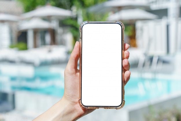 Mano de mujer sosteniendo teléfono móvil de pantalla en blanco junto a la piscina.