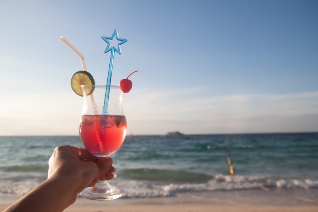 Mano de mujer sosteniendo un vaso de bebida de verano de color rojo con playa