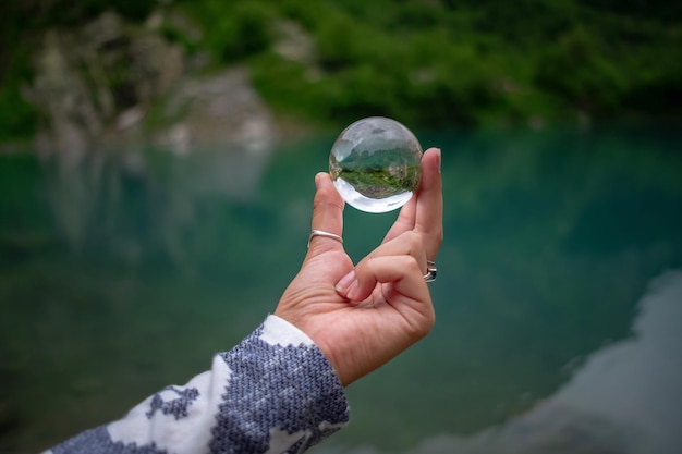 La mano de una mujer sostiene una bola de cristal esférica contra el fondo de un hermoso lago de montaña