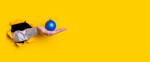 La mano de la mujer sostiene una bola de Navidad decorativa azul sobre un fondo amarillo Banner