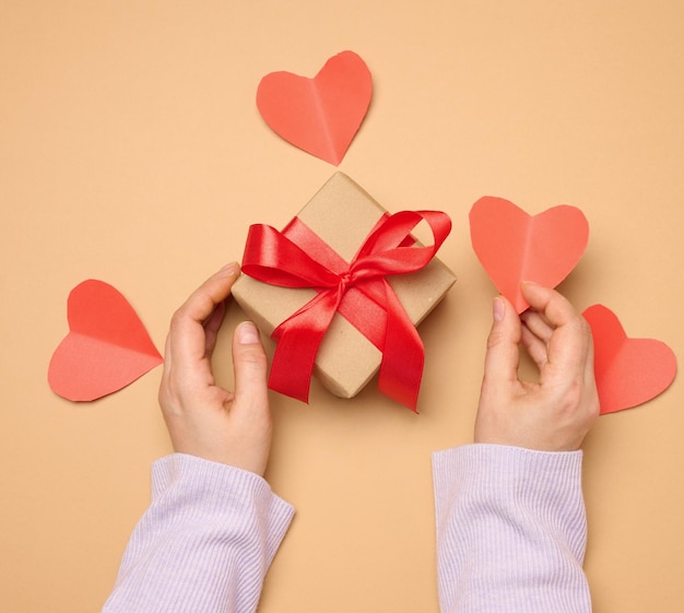 La mano de una mujer sostiene una caja de regalos envuelta en una cinta de seda roja sobre un fondo beige