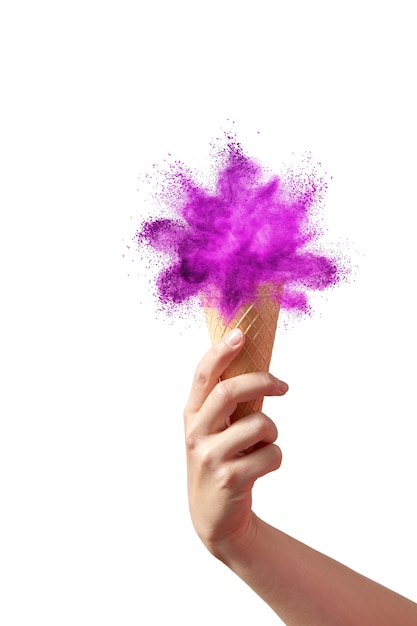 La mano de la mujer sostiene el cono de galleta con salpicaduras de polvo abstracto coloreado como un helado dulce en una pared blanca, copie el espacio.