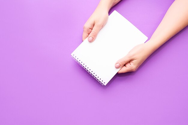 La mano de una mujer sostiene un cuaderno con hojas blancas sobre un fondo claro. Un lugar para el texto, una vista desde arriba. Endecha plana