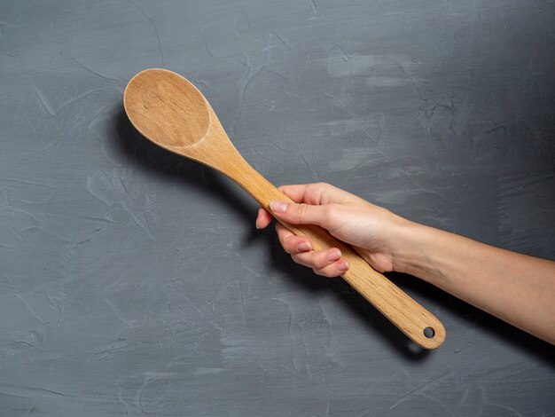 La mano de una mujer sostiene una cuchara de madera grande sobre un fondo de textura gris. Herramientas para cocinar alimentos.