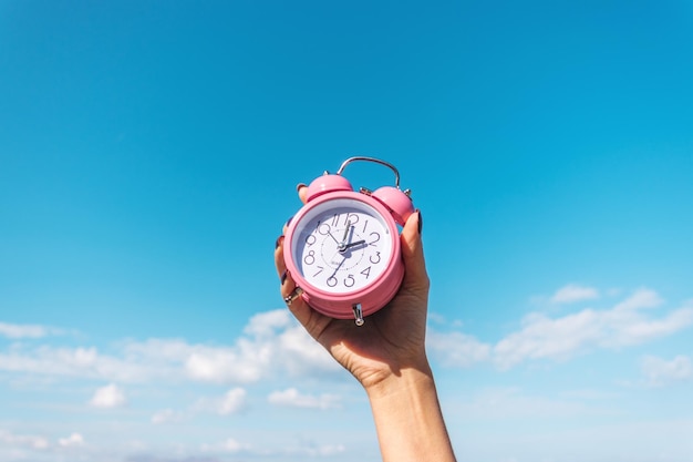 La mano de la mujer sostiene el despertador rosa en el fondo del cielo. Concepto de vacaciones de verano.