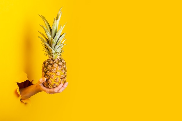 La mano de la mujer sostiene una piña entera sobre un fondo amarillo roto.