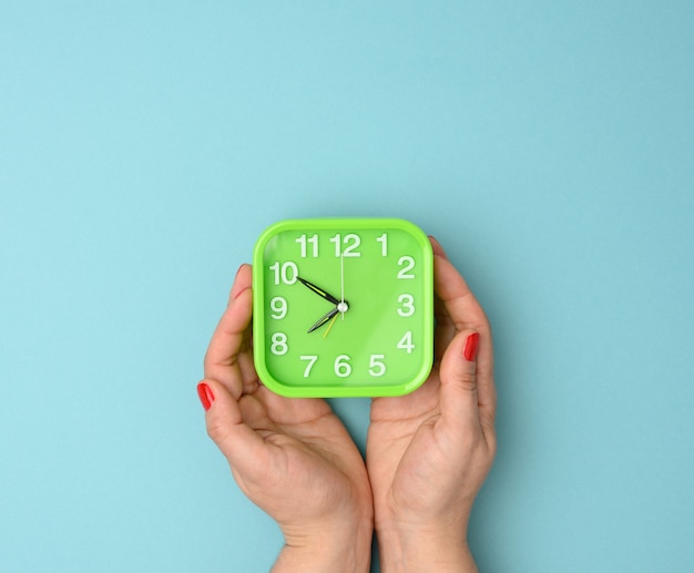 La mano de la mujer sostiene un reloj despertador cuadrado verde, la hora es de diez a ocho de la mañana. Concepto de madrugada, fondo azul