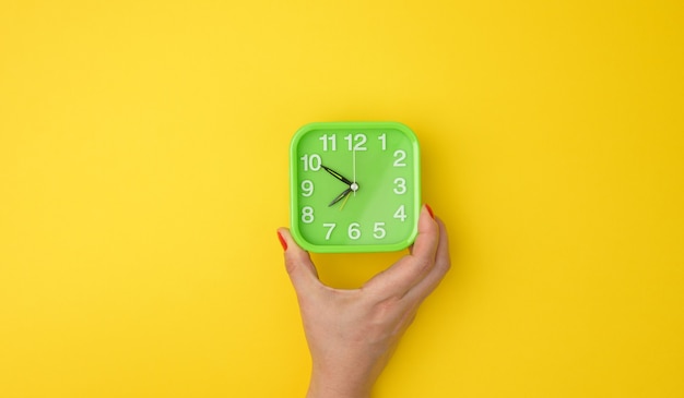 La mano de la mujer sostiene un reloj despertador cuadrado verde, el tiempo es de diez a ocho de la mañana