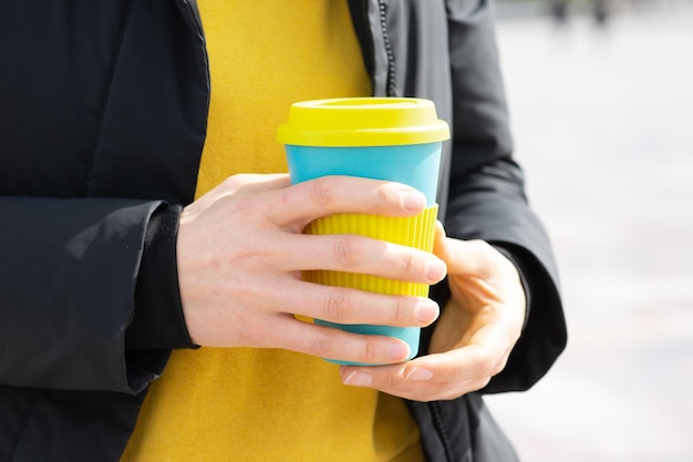 La mano de una mujer sostiene una taza de café ecológica en el fondo de una calle de la ciudad, toma café. Desayuno sobre la marcha.