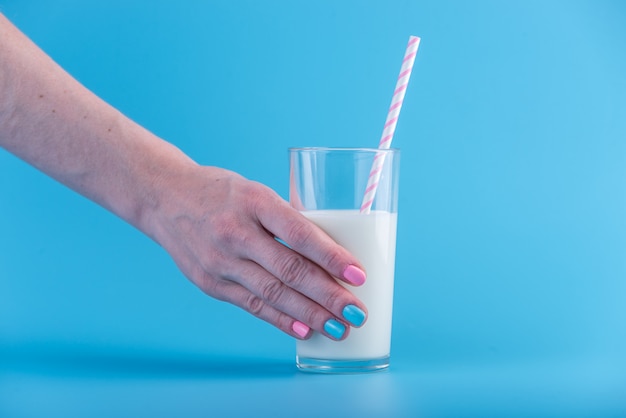 La mano de la mujer sostiene un vaso de leche fresca con una pajita sobre un fondo azul. Concepto de productos lácteos saludables con calcio.