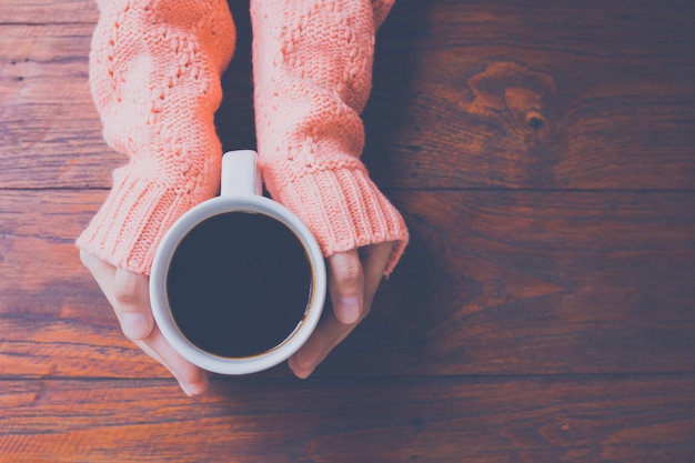 Mano de la mujer en el suéter caliente que sostiene una taza de café en un fondo de madera de la tabla