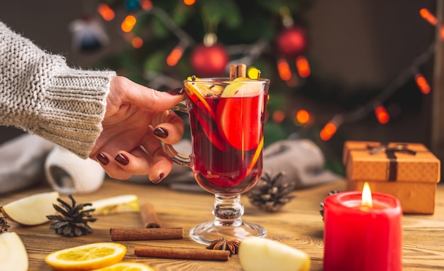 La mano de una mujer en un suéter caliente sostiene una taza de vino caliente aromático caliente contra el árbol de Navidad