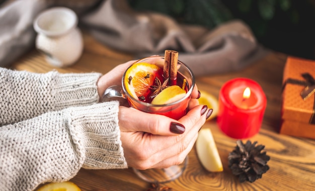 La mano de una mujer en un suéter caliente sostiene una taza de vino caliente aromático caliente contra el árbol de Navidad