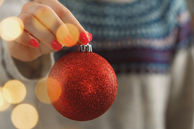 Mano de una mujer en suéter festivo sosteniendo adornos navideños con luces borrosas de cerca