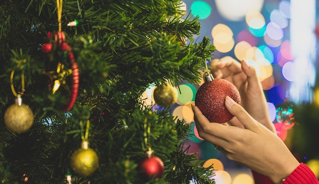 Mano de mujer en suéter de santa colgando coloridos adornos encantadores y bola en el árbol de Navidad para decorar la hermosa fiesta de celebración en la noche feliz. Agregue un poco de ruido para adaptarse a la imagen de estilo vintage.