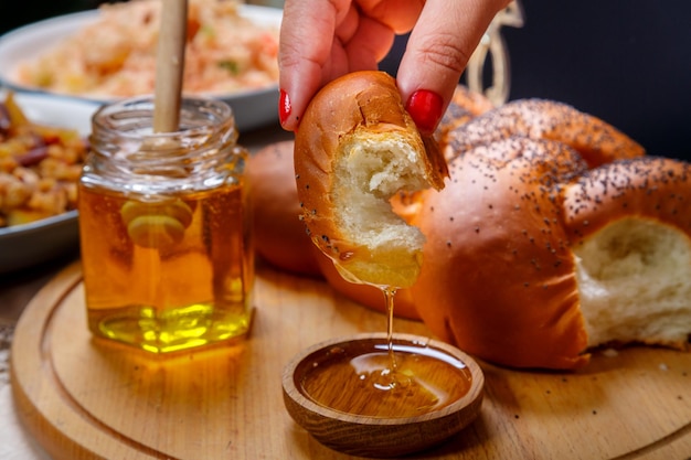 La mano de una mujer sumerge un trozo de jalá en miel en honor a la celebración de Rosh Hashaná cerca de la mesa miel y jalá y comida tradicional