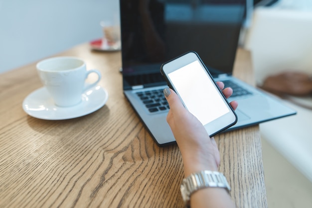 Mano de mujer con teléfono en mano contra laptop y café