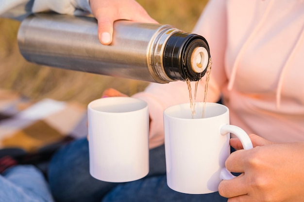 La mano de una mujer con un termo vierte té caliente en una taza blanca, primer plano. Amigas adultas en un picnic de otoño