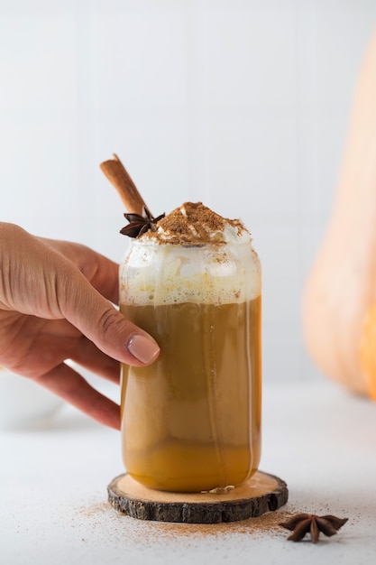 La mano de una mujer toma una taza de cristal de delicioso café de calabaza con crema batida y una ramita de canela