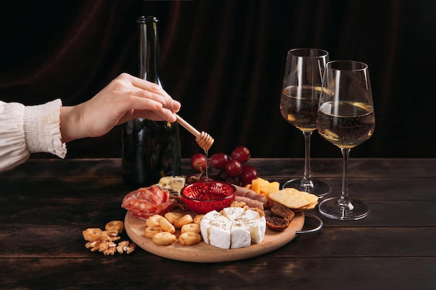 La mano de la mujer toma un trozo de queso del tablero de aperitivos con una variedad de queso, carne, salchicha, roseta, uva y galletas