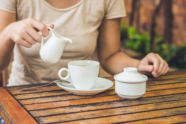 Mano de mujer vertiendo leche en una taza de café blanco en la cafetería al aire libre