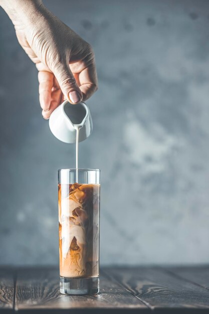 La mano de las mujeres está vertiendo crema agria casera de un frasco pequeño a un vaso con café frío y hielo Bebida fría de verano en una mesa de madera oscura y fondo gris con espacio para copiar