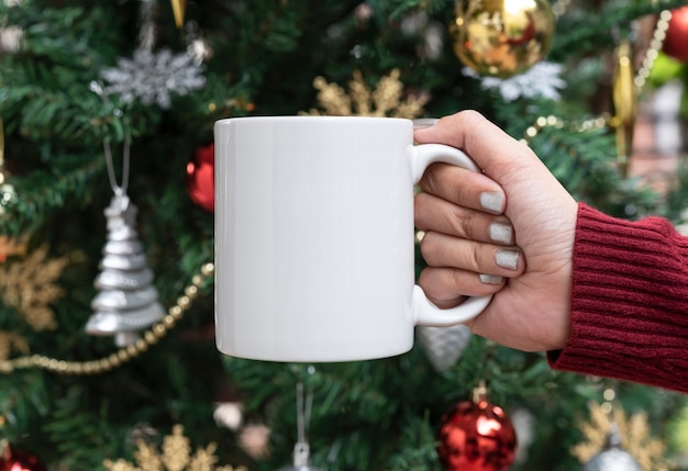 Mano de las mujeres que sostiene la taza de café de cerámica blanca en fondo del árbol de navidad. maqueta para mensajes de texto publicitarios creativos o contenido promocional.