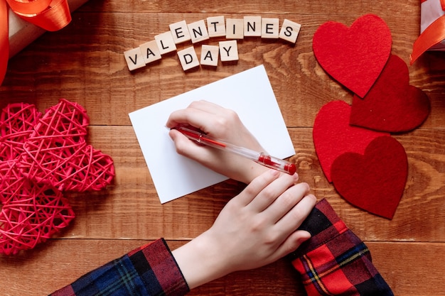 Foto la mano de la niña escribe una carta de amor. día de san valentín. tarjeta de felicitación hecha a mano con un corazón rojo en forma de figura. el 14 de febrero es una celebración de celebración. vista desde arriba