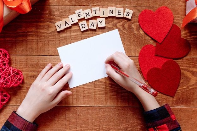 La mano de la niña escribe una carta de amor. Día de San Valentín. Tarjeta de felicitación hecha a mano con un corazón rojo en forma de figura. El 14 de febrero es una celebración de celebración. Vista desde arriba