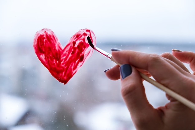 Mano de una niña con un pincel pintando corazón rojo en una ventana, quedarse en casa, ocio en cuarentena, estemos todos bien, día de San Valentín, amor, romance