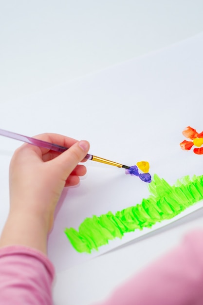 Mano de niño dibujando flores sobre la hierba verde con pincel de acuarelas sobre papel blanco.