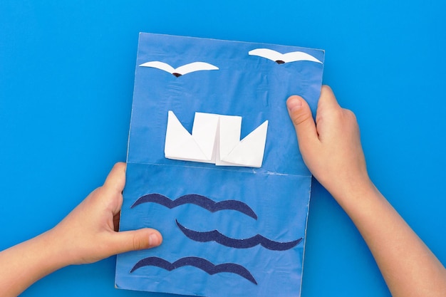 Mano de niño plano con tarjeta de felicitación artesanal de papel con mar de barco y gaviotas presentes para el padre sd