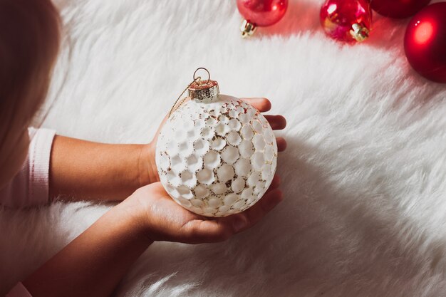 La mano de los niños sostiene una bola blanca de Navidad contra una manta de lana suave blanca