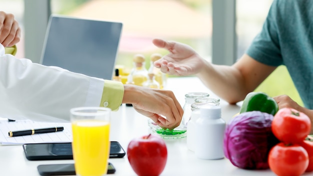 Mano del nutricionista agarrando píldoras verdes de la botella en la mesa para mostrárselas al paciente. Frutas y verduras, jugo, computadora portátil, teléfono móvil en la mesa de la oficina del nutricionista.