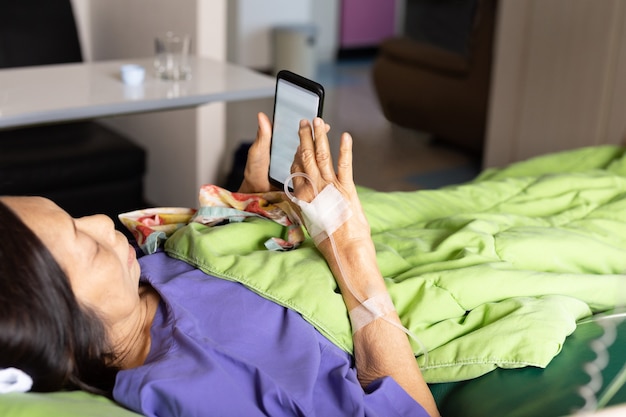 Mano del paciente mayor con inyección de solución salina acostado con teléfono celular en el hospital