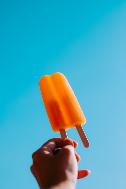 Mano con paleta de hielo naranja en verano soleado cielo azul brillante.
