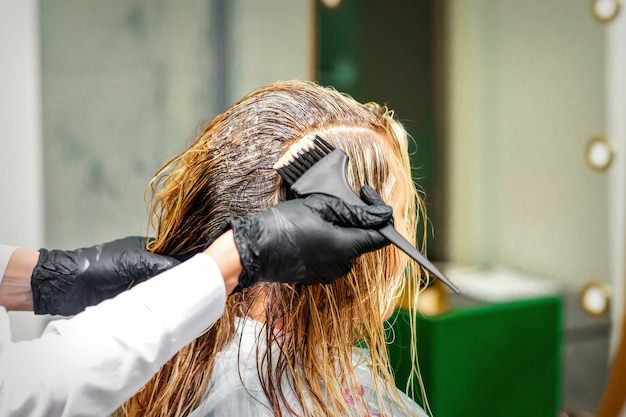 Mano de un peluquero en guantes negros aplicando tinte para el cabello femenino en un salón de belleza