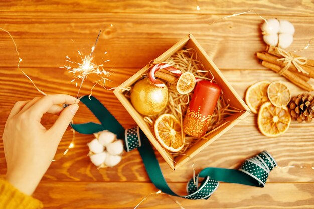 La mano de las personas sostiene la bengala sobre la mesa de madera con caja de regalos de navidad con vela adornada bola dorada ...