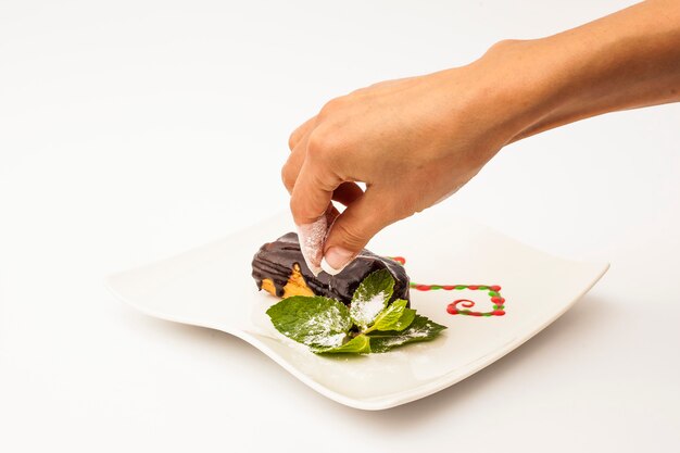 Mano poniendo un poco de azúcar en la torta de Eclair con chocolate en un plato blanco con una menta sobre un fondo blanco.