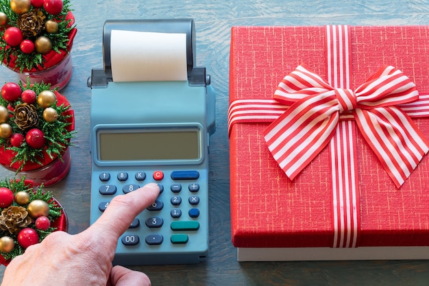 Una mano presionando los botones de la caja registradora junto a una caja de regalo roja con una cinta y pequeños árboles de navidad con conos. cálculo del monto del obsequio. comprar regalos para navidad