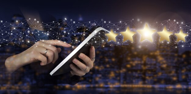 Mano que señala el símbolo de cinco estrellas para aumentar la calificación de la empresa. Tableta blanca táctil de mano con holograma digital de cinco estrellas firmar sobre fondo borroso oscuro de la ciudad. Concepto de evaluación de servicio de producto.