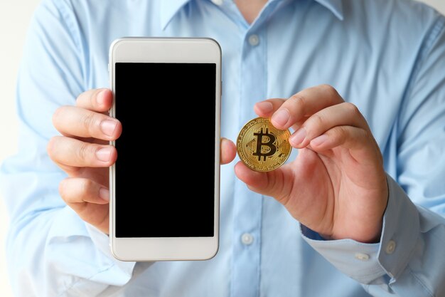 Mano que sostiene bitcoin y teléfono inteligente con fondo de pantalla en blanco, simulacro, criptomoneda