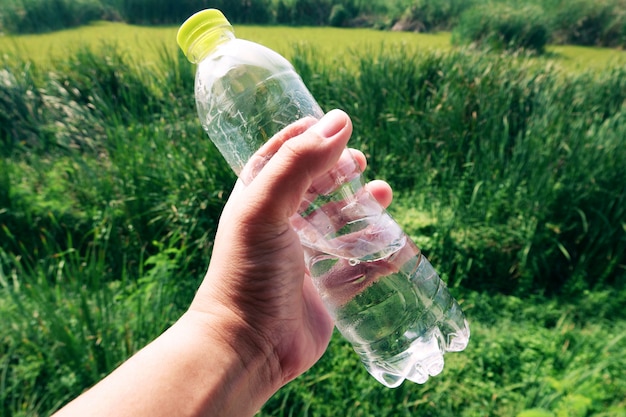 Mano que sostiene la botella de agua plástica sobre fondo verde jardín borrosa