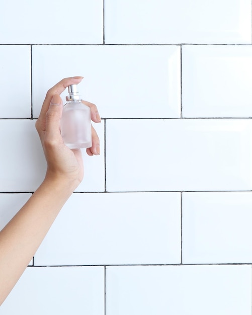 Mano que sostiene la botella de vidrio de perfume sobre fondo de mosaico minimalista blanco. Botella de spray Eau de Toilette.