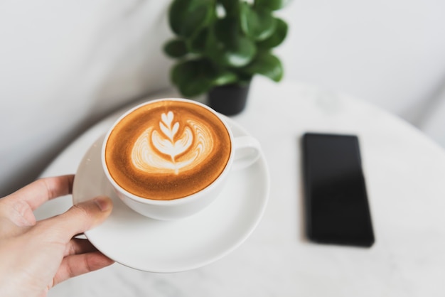 Mano que sostiene el café con leche o capuchino con espuma espumosa, vista superior de la taza de café en la cafetería.